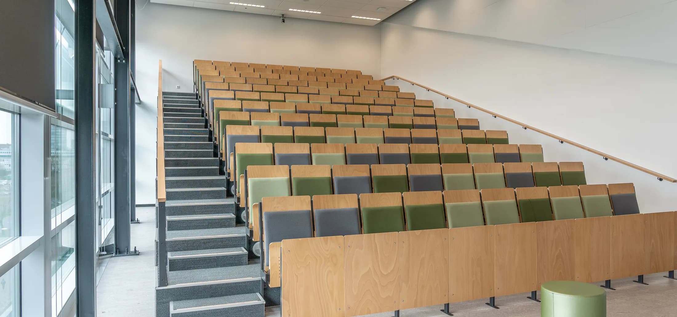 Tomt auditorium med tomme rækker af træsæder i grønt og gråt, dagslys gennem en glasfacade til venstre