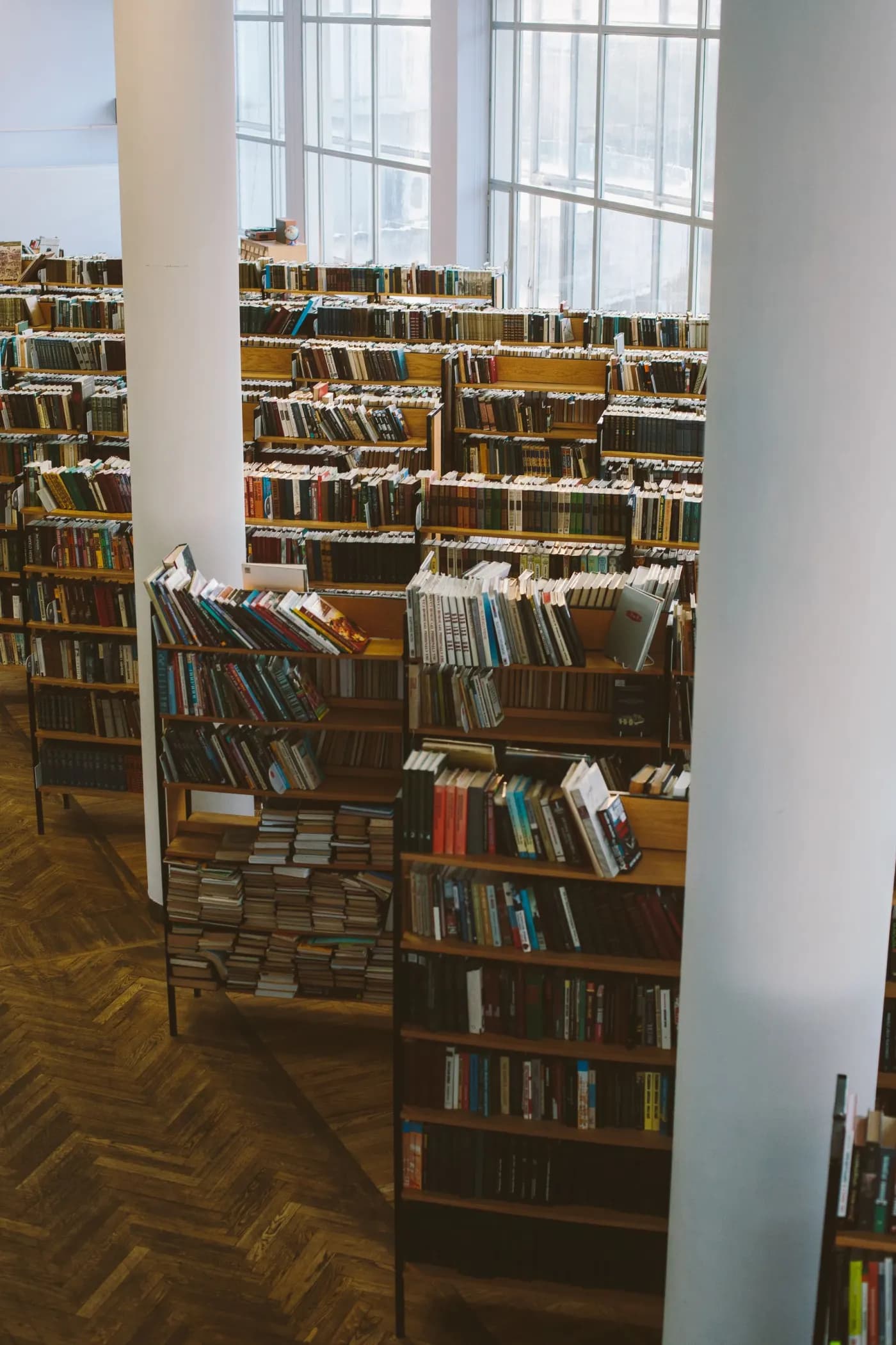 Et biblioteks læsesal set mellem to hvide søjler, træreoler fyldt med bøger, høje vinduer og parketgulv, ingen mennesker